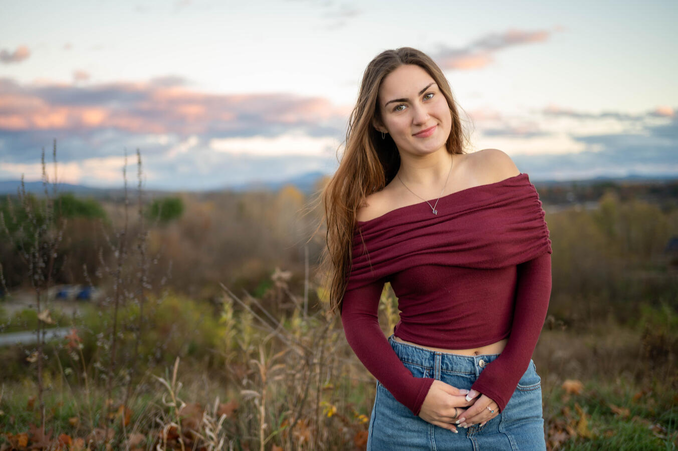A young woman poses for a senior class portrait in colchester, vermont at saint michael's college. She wears a red top in front of a sunset background. camel's hump mountain can be seen in the distance. Photo by Paul Detzer.