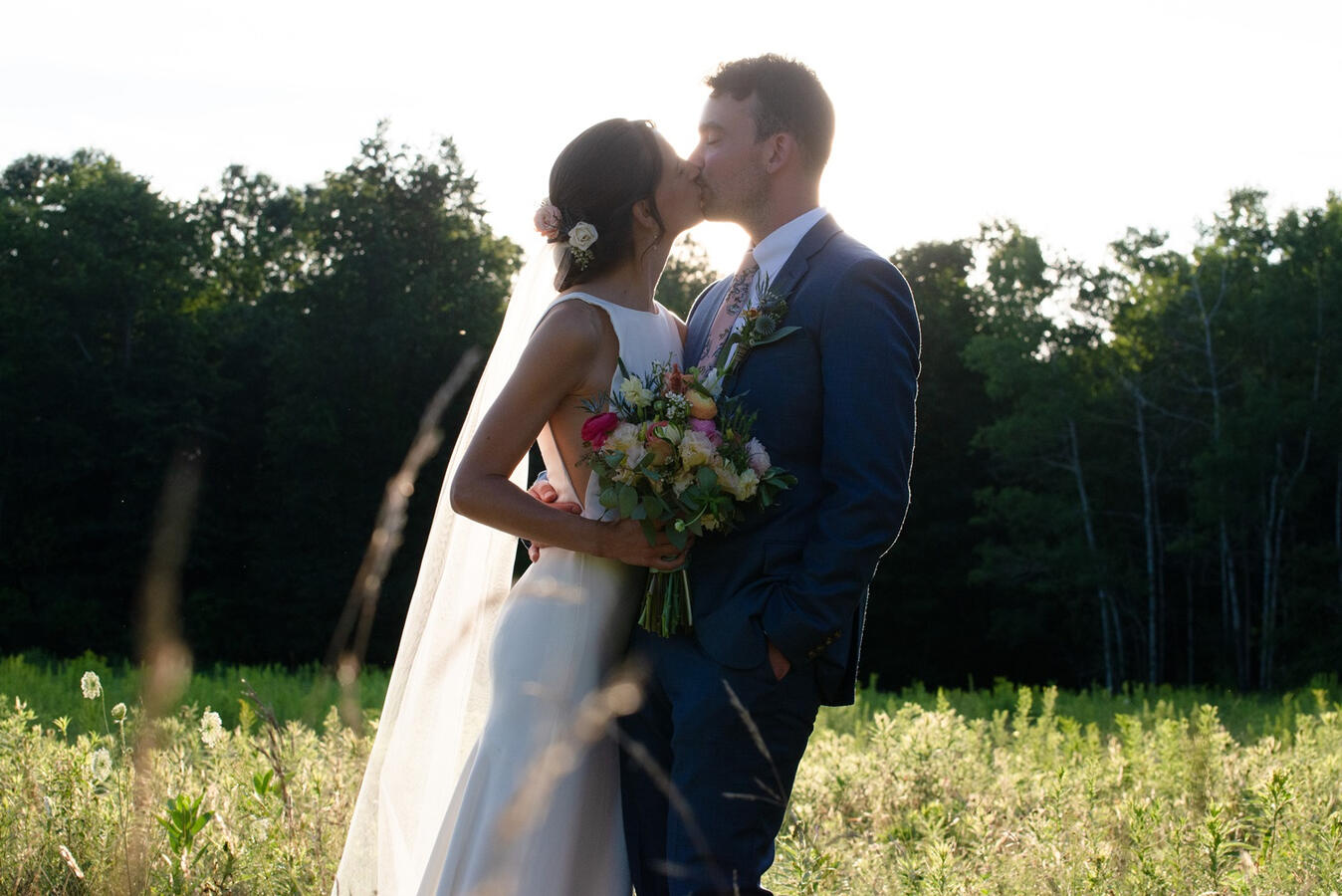 A bride and groom kiss in the sunset, silhouetted by the light